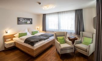 A modern hotel room with a large double bed, two comfortable armchairs, and large windows. The decor is in warm, bright colors, perfect for relaxing.