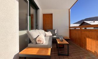 A cozy balcony with a comfortable seating area and a wooden table. In the background, snow-covered mountains and a blue sky can be seen.
