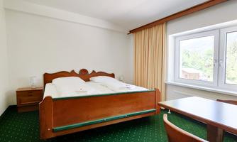 A simple room with a double bed and a table. Large windows let in a lot of light and the carpet is green.