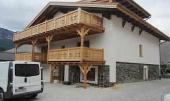 A modern house with wooden cladding and two balconies. In front of the house, there is a white utility vehicle on a paved area.