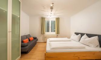 A cozy room with a double bed and a sofa bed. Wooden floor and large windows create a bright atmosphere.
