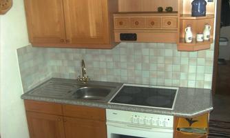 A modern kitchen unit with wooden cabinets and a light tiled backsplash. The equipment includes a cooktop, an oven, and a sink.