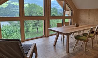A bright, modern room with large windows that offer a view of green trees and mountains. The room is furnished with a wooden table and chairs.