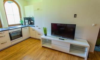 A modern kitchen with a TV and a white TV cabinet. The floor is made of wood and there is plenty of natural light.