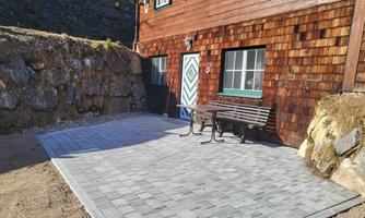 A cozy terrace made of gray paving stones in front of a wooden house. A bench is placed at a table, surrounded by a stone wall.