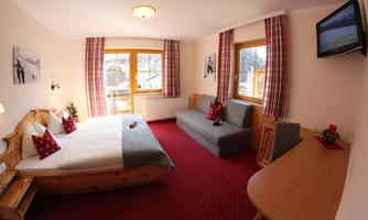 A cozy hotel room with two beds and a sofa. Large windows provide plenty of light, and the decor is rustic with red accents.