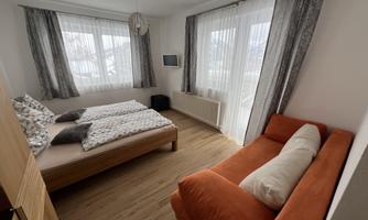 A modern bedroom with two beds and a cozy orange sofa. Large windows let in plenty of daylight.