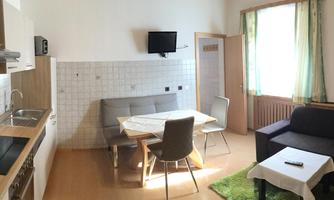A modern kitchen with a dining area and a cozy seating corner. Large windows provide plenty of daylight.