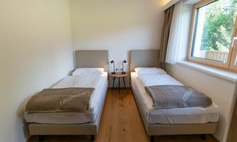 A simple bedroom with two single beds and a small table in the middle. The room has large windows that let in daylight.