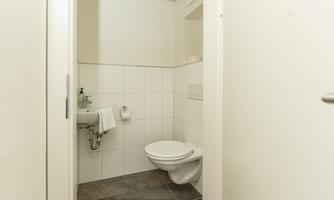 A modern bathroom with white tiles and a sink. The toilet is tidy and the room is well-lit.