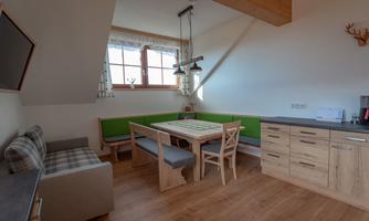 A bright and cozy kitchen with wood veneer and a green upholstered bench. The table sits in the middle, surrounded by chairs, and large windows provide plenty of daylight.