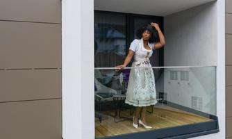 A woman in a traditional dress stands on a balcony and looks outside. The room is designed in a modern style, and the weather seems pleasant.