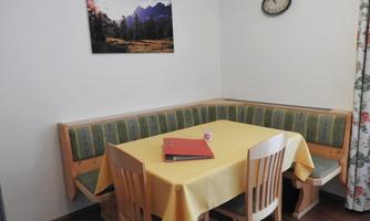 A cozy dining area with a bench and a table, dressed with a yellow tablecloth. A picture of mountains hangs on the wall, and the clock shows the time.