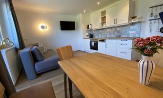A cozy kitchen with a wooden dining table and a modern living room corner. The decoration includes a vase with flowers and a clear, bright interior.