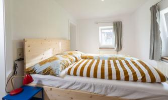 A bright bedroom with a large bed and colorful pillows. The walls are plain and there is a window with curtains.
