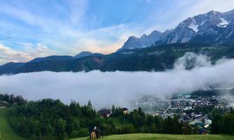 A picturesque landscape with mountains in the background and a deep layer of fog over a village. The sky is blue with a few clouds and the meadows are green.