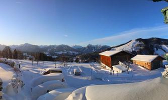 A snowy landscape with mountains in the background. The sun shines over the snow-covered roofs and meadows.