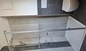 A modern bathroom with a transparent shower cubicle and a simple toilet. The walls are tiled and kept in neutral colors.
