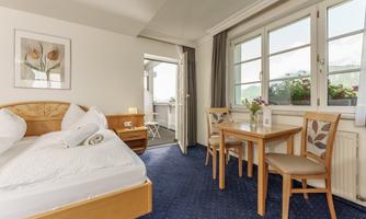 A cozy bedroom with a double bed and a small table. Large windows let in plenty of daylight and offer a view of the outdoor area.