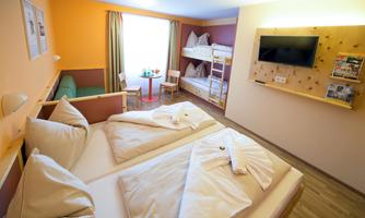 A bright hotel room with two beds and a bunk bed. There is a small table and a television on the wall.
