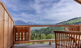 A beautiful balcony with a wooden railing and a view of the mountains. In the background, green hills and a clear sky can be seen.
