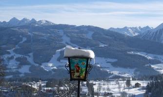 A winter landscape with snow-covered mountains and a clear sky. In the foreground stands a lit lantern with snow on top.