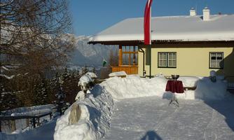 A wintry scene with a snow-covered exterior in front of a house. In the background, mountains and a blue sky are visible.