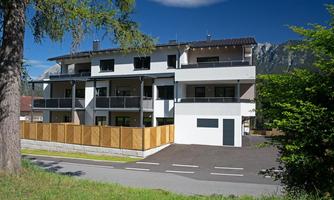 A modern residential building with several balconies and a wooden fence. In the background, green trees and mountains are visible.