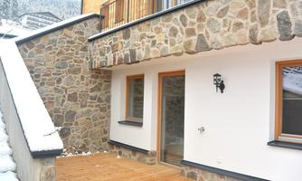 A modern terrace with a wooden floor and a stone wall. The area is covered with snow, and large windows let light in.