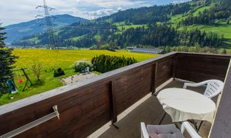 A balcony with a table and two chairs offers a view of green meadows and mountains. In the background, power lines and a beautiful hilly landscape are visible.