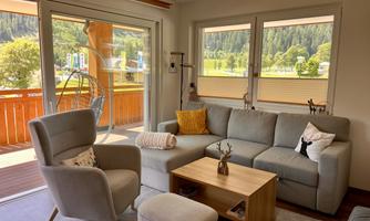 A bright living room with a cozy sofa corner and large windows overlooking a green landscape. The room is modern and invitingly furnished.