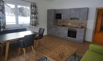A modern kitchen with a dining area and a cozy sofa. The room design is bright and friendly with large windows.