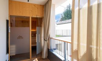 A modern balcony with bright curtains and a wooden chair. In the background, a room with wooden paneling can be seen.
