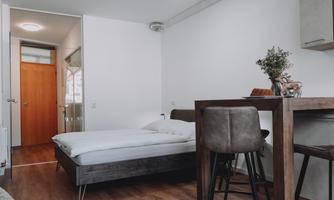 A modern room with a bed and a small kitchenette. There is a table with chairs and plenty of natural light.