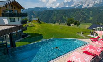 A modern hotel with a pool and red sun loungers. In the background, green meadows and majestic mountains stretch under a blue sky.