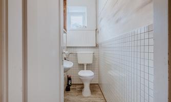 A modern bathroom with wooden floors and white walls. There is a toilet and a narrow hallway that leads to the room.