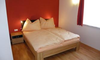 A cozy bedroom with a double bed and two pillows. The walls are in warm colors, and a window lets in daylight.
