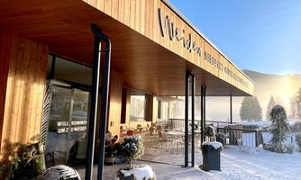 A modern wooden building with large windows and an inviting terrace. The surroundings are covered in snow and there seems to be a calm, clear winter atmosphere.