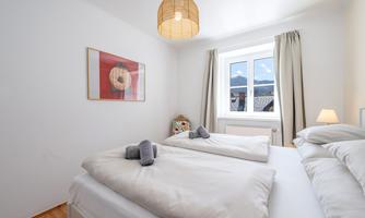 A cozy bedroom with two single beds and a bright window front. The walls are kept in a simple white, and a lamp provides warm light.