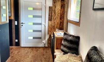 A bright entrance area with wooden flooring and a modern glass door. Natural wood details are on the wall and a cozy seating area with furs.