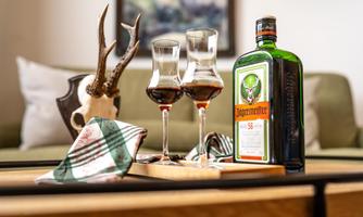A bottle of Jägermeister is standing on a table next to two glasses with a dark drink. In the background, a cozy living environment can be seen.