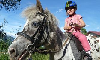 A little girl rides a spotted pony. She is wearing a helmet and smiles at the camera while the surroundings are green and sunny.