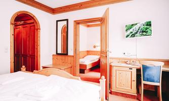 A bright room with wooden furniture and a double bed. In the background, another room with a bed and a small desk is visible.