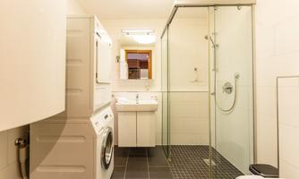 A modern bathroom with a shower and a washer-dryer combination unit. The walls are bright tiled and the floor features a dark tile pattern.