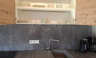 A modern kitchen with a gray countertop and light wood cabinets. Dishes are on a shelf above the sink.