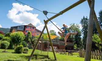 A beautiful playground with a large swing, where two children are happily playing. In the background, you can see a colorful building and many green trees.