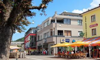 A lively city street with cafés and colorful sun umbrellas. In the background, modernly designed buildings and a blue sky can be seen.