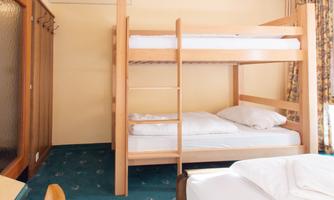 A simple room with a loft bed and another bed in the foreground. The walls are painted in a light shade, and the floor is carpeted.