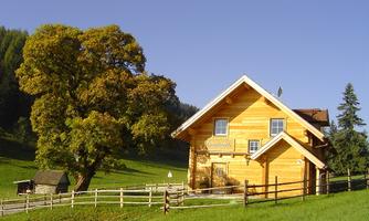 A cozy wooden house stands amidst green meadows. A large tree and a fence surround the property in a picturesque landscape.