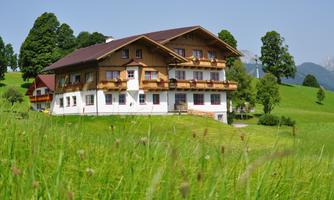 A charming building in alpine style, surrounded by green meadows and trees. In the background, gentle hills and a clear sky can be seen.